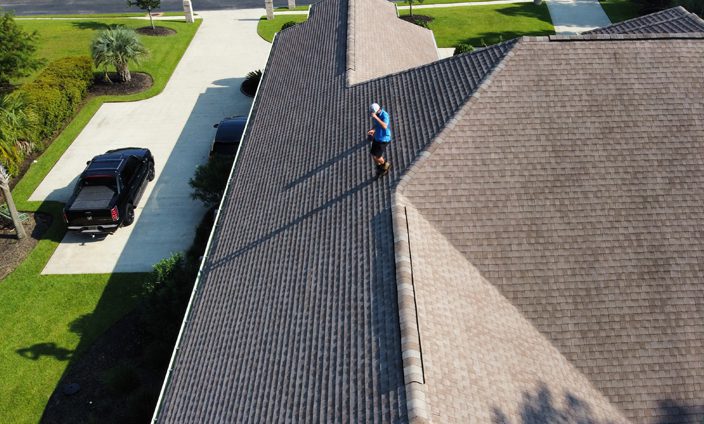 Weatherguard Roofing & Restoration | A Weatherguard Roofing and Restoration team member inspecting a roof of a house with a black truck in the driveway and a yard lined with shrubs and a palm tree